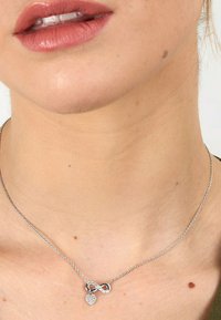 Silberne Halskette mit einem Unendlichkeitssymbol und einem kleinen Herzanhänger, geschmückt mit klaren Steinen, aufgehängt an einer zarten Kette.