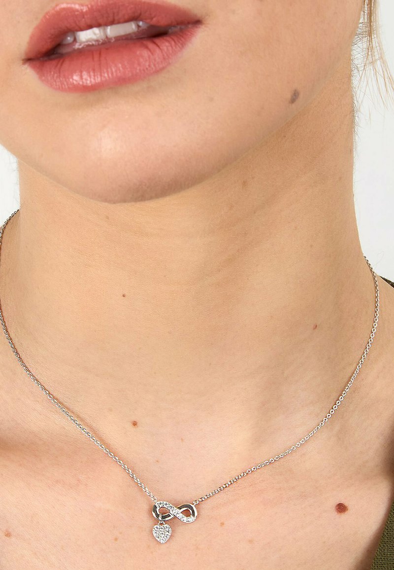 Silberne Halskette mit einem Unendlichkeitssymbol und einem kleinen Herzanhänger, geschmückt mit klaren Steinen, aufgehängt an einer zarten Kette.