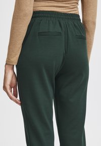 Close-up van een persoon die een donkergroene elastische broek en een beige langarmige top draagt, met zichtbare achterzakken en een hand rustend aan de zijkant.