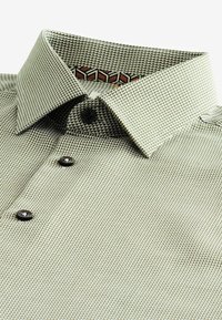 Chemise habillée pour hommes avec col, présentant un petit motif géométrique vert et blanc et des boutons noirs, soigneusement pliée sur un fond blanc.