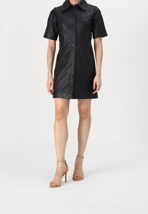 Femme portant une robe en cuir noire à manches courtes avec boutons et des escarpins beiges à bride à la cheville, debout sur un sol blanc.