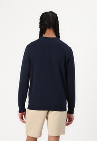 Personne aux longs cheveux tressés vue de dos, portant une chemise à manches longues bleu marine et un short beige sur fond blanc uni.