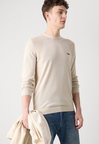 Beige neulepusero, jossa on ribbinelementtejä olkapäissä ja hihoissa. Malli pitää kädessään kreikkalaista takkia ja on pukeutunut sinisiin farkkuihin. Logo näkyy rinnassa.