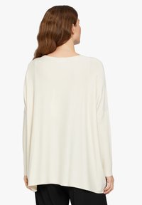 Beigefarbener Pullover mit lockerer Passform, langen Ärmeln und rundem Ausschnitt. Weiche Textur, mit einem längeren Rückensaum und seitlichen Schlitzen.