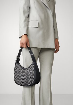 Sac à main en cuir noir avec des découpes circulaires, forme courbée et bandoulière cloutée, tenu par une personne en costume gris clair.