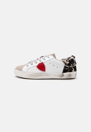 Baskets basses blanches avec bout beige, empiècement latéral rouge, lacets gris et détail en fourrure noire et blanche tachetée au talon et au col.