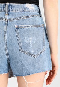 Pantalones cortos de mezclilla azul claro con dobladillo deshilachado, detalles desgastados y un bolsillo trasero con un patrón en forma de T cosido con hilo más claro.