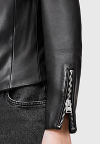 Giacca in pelle nera con design fitted, dettagli con maniche zip e texture liscia. Abbinata a jeans scuri.