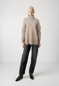 Beige gestrickter Rollkragenpullover mit lockerer Passform und geripptem Saum, kombiniert mit lässigen schwarzen Jeans und glänzenden schwarzen Stiefeletten.