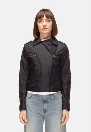 Donna che indossa una giacca nera asimmetrica con zip sopra una camicia bianca abbinata a jeans a vita alta color azzurro chiaro, in piedi davanti a uno sfondo uniforme.