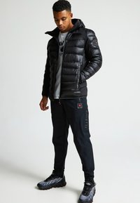 Čierna nafukovacia bunda s kapucňou, so zipsom a prešívaným dizajnom. V kombinácii s čiernymi cargo nohavicami a sivými športovými topánkami.