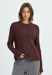 Maglione lavorato a maglia in un profondo bordeaux con una texture a coste, collo rotondo e maniche allungate. Abbinato a pantaloni grigi sartoriali.