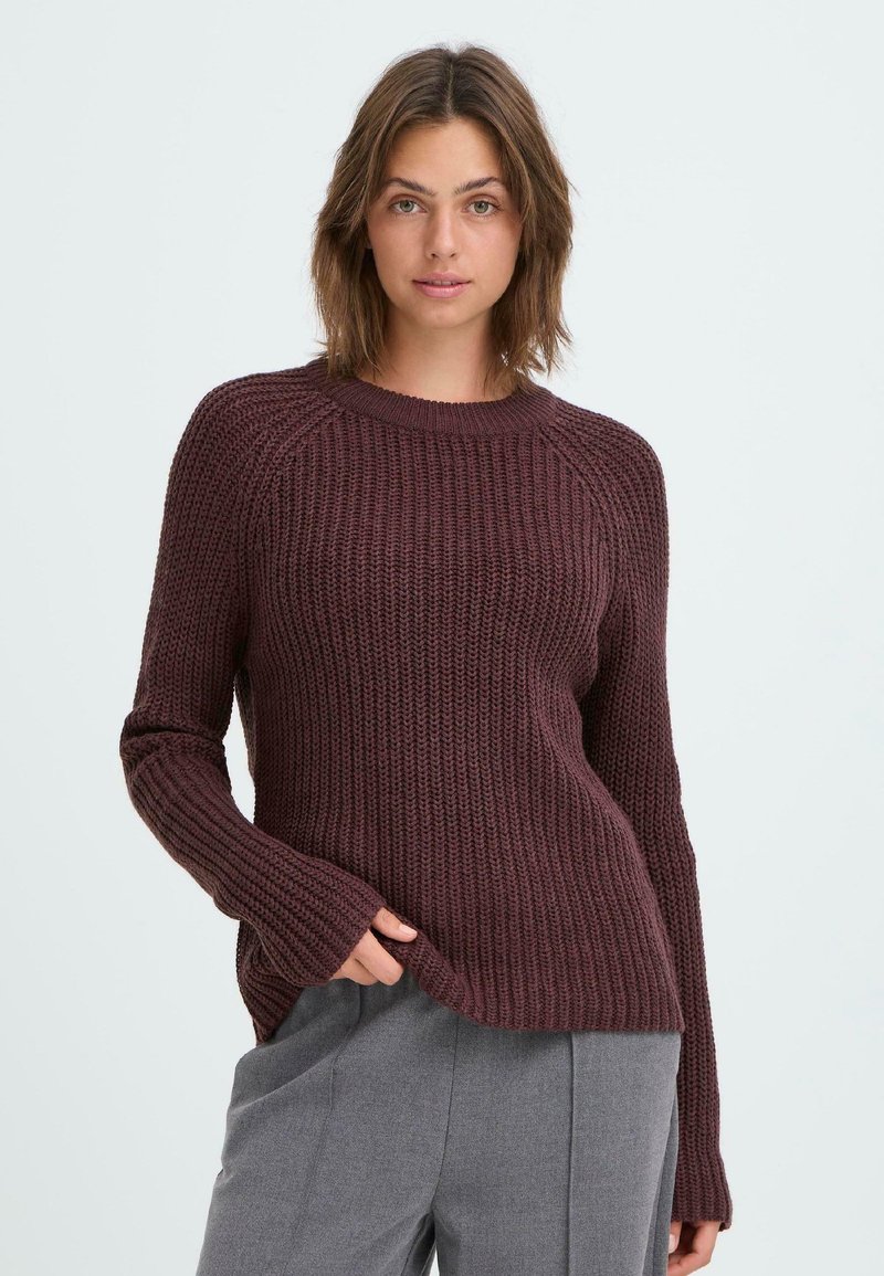 Maglione lavorato a maglia in un profondo bordeaux con una texture a coste, collo rotondo e maniche allungate. Abbinato a pantaloni grigi sartoriali.