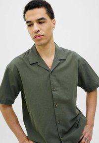 Chemise à manches courtes, boutonnée, en tissu vert olive. Dispose d'un col de type camp et de boutons marron. Texture douce avec une coupe décontractée.