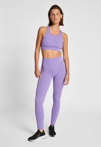 Kvinna i lila ribbad sport-bh och högmidjade leggings, bär svart sportskor, stående mot en enkel ljus bakgrund.