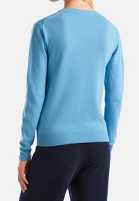 Nainen, jolla on vaaleansininen pitkähihainen neule ja tummat housut, kuvattuna selkäpuolelta valkoista taustaa vasten.