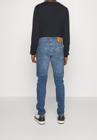 Blå denimjeans med rak bendesign, subtil blekning och en brun läderlapp vid midjan, i kombination med en svart långärmad skjorta.