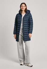 Manteau matelassé bleu marine avec capuche, doté d'une fermeture éclair à l'avant, d'un design matelassé et d'une coupe longue. Associé à un jean large gris clair et des baskets.