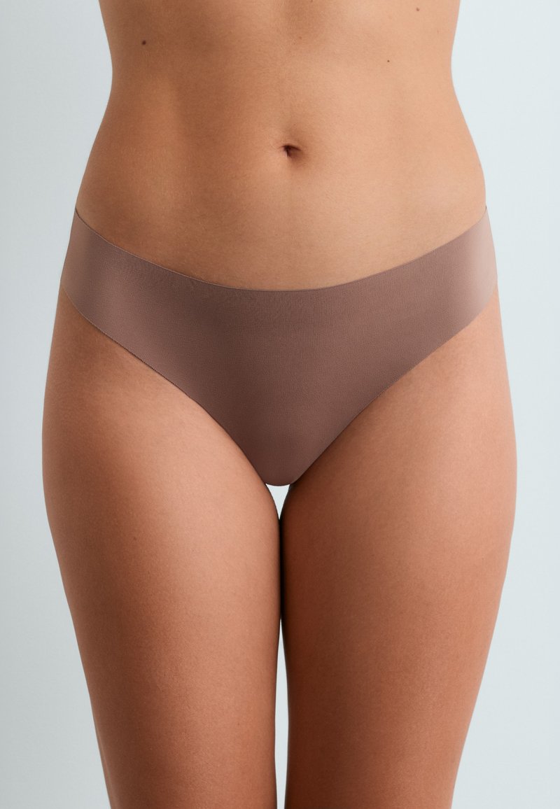 Femme portant des sous-vêtements marron sans couture, montrant la partie médiane et le haut des cuisses sur un fond neutre.