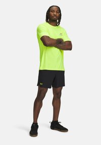 Neon-gelbes Sportshirt mit kurzen Ärmeln und Belüftungsdetails, kombiniert mit schwarzen Shorts und schwarzen Sportschuhen. Logo-Akzente vorhanden.