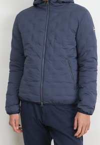 Man draagt een doorgestikte marineblauwe jas met ritssluitingen aan de zijzakken en bijpassende marineblauwe broek, staande tegen een effen achtergrond.