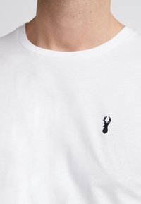 Hvid bomulds t-shirt med rund halsudskæring. Har et lille sort broderet hjort-logo på venstre bryst. Glat tekstur.