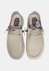 Chaussures slip-on en toile beige avec une finition texturée, des lacets élastiques et une doublure à motifs dans des couleurs claires. Logo visible sur les côtés.