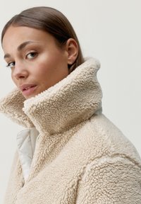 Beżowa kurtka z sztucznego futra z wysokim kołnierzem i błyszczącym zapięciem na zamek. Miękka, teksturowana tkanina zewnętrzna oraz gładka podszewka.