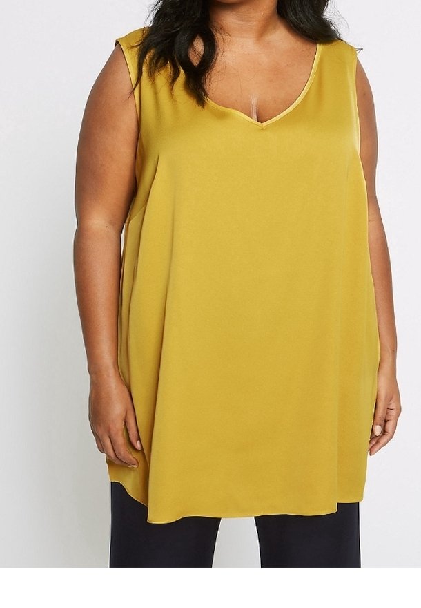 Femme portant un haut tunique sans manches jaune moutarde à col en V, associé à un pantalon noir, sur un fond clair uni.