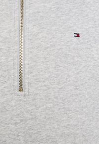 Sweat-shirt gris clair avec une fermeture éclair quart de zip dorée. Présente un petit logo rouge, blanc et bleu sur la poitrine. Tissu doux et texturé.