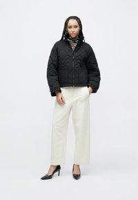 Czarna pikowana kurtka z wysokim kołnierzem, zamkiem z przodu i dwoma bocznymi kieszeniami, w połączeniu z kremowymi spodniami o szerokich nogawkach i czarnymi szpiczastymi szpilkami.