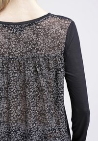 Zwarte lange mouwen top met een doorschijnend achterpaneel met witte bloemmotieven. De onderrug heeft een geplooid ontwerp voor extra detail.
