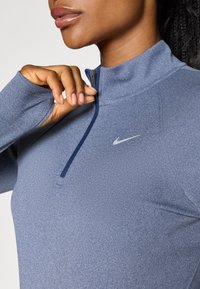 Blå atletisk jakke med glidelås laget av teksturert stoff. Har høy krage, front med halv-glidelås, og en liten hvit Nike-logo på brystet.