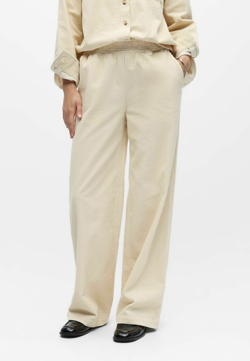 Pantalones beige de pierna ancha con superficie texturizada, cintura elástica, bolsillos laterales y ajuste holgado, combinados con una parte superior a juego.
