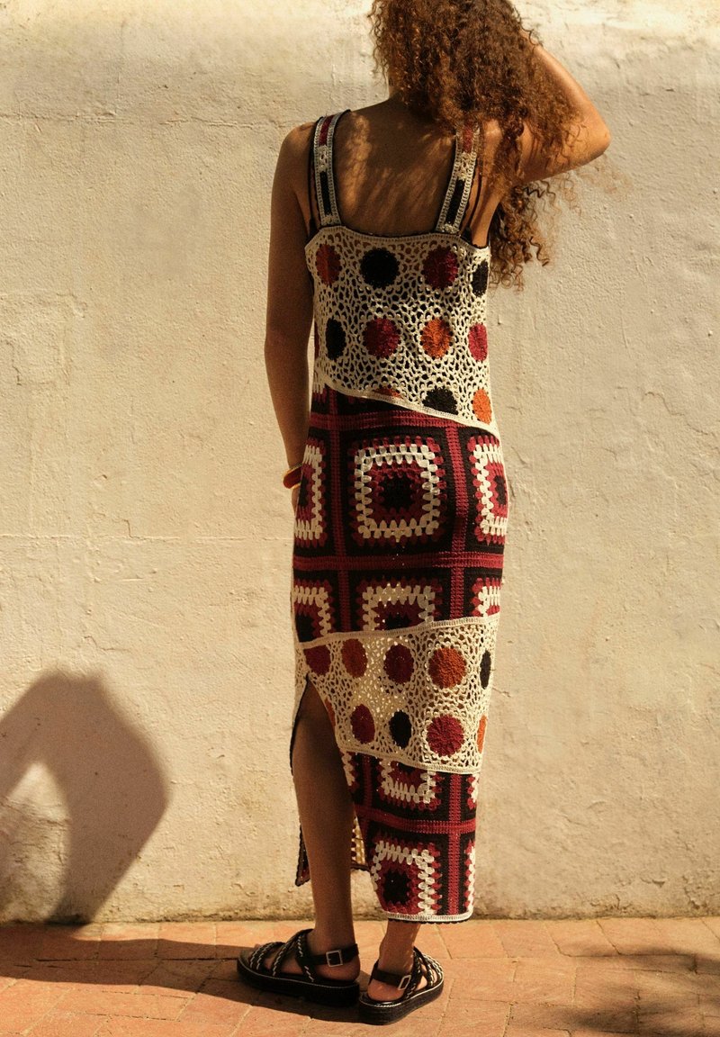 Häkelkleid mit einem gemusterten Design in Rot-, Schwarz- und Creme-Tönen; verfügt über seitliche Schlitze und breite Träger. Wird mit dunklen Sandalen getragen.