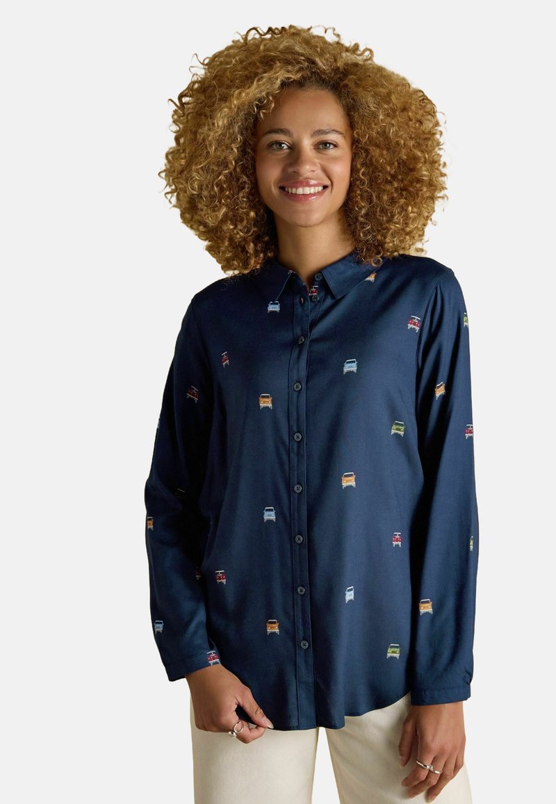 Woman with curly hair wearing a navy blue button-up shirt with small colorful car patterns and light-colored pants, smiling.
