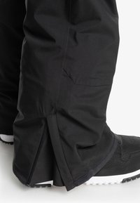 Pantalones impermeables negros con una textura suave, que presentan una apertura lateral con cremallera y un dobladillo ligeramente acampanado, combinados con zapatillas deportivas negras.