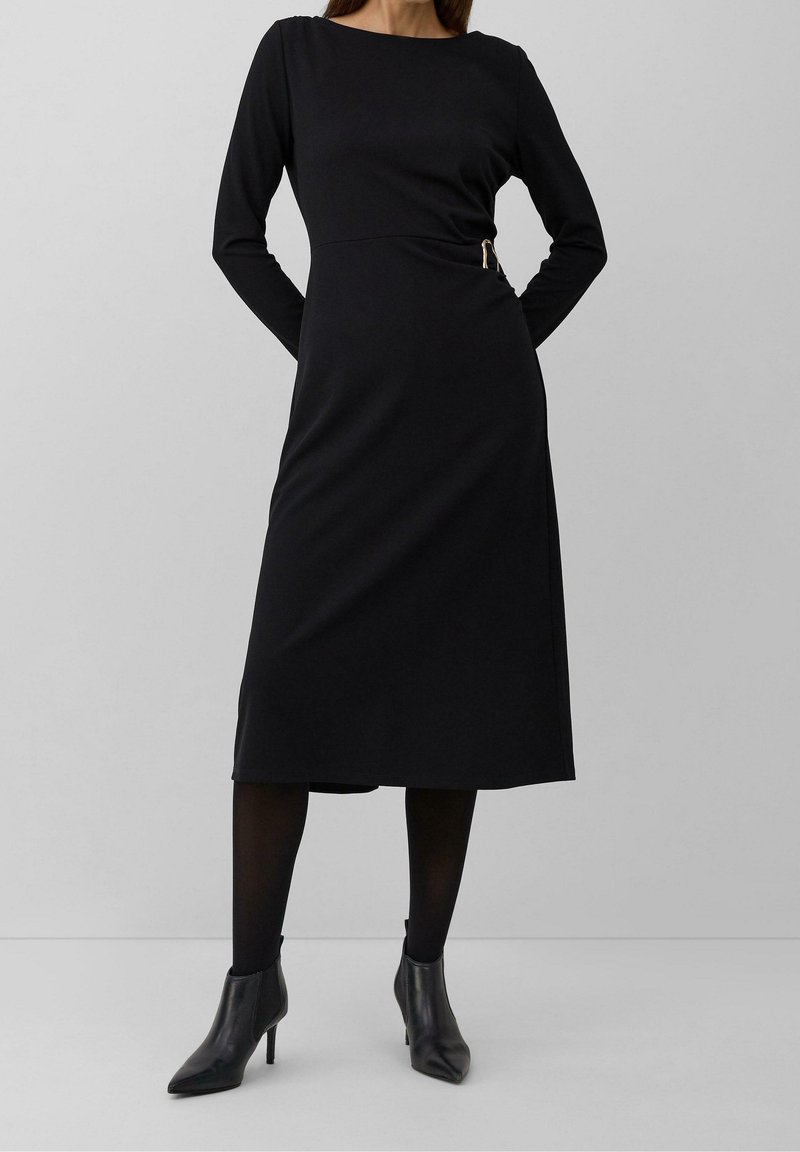Femme portant une robe midi noire à manches longues avec un détail de boucle sur le côté, des collants noirs et des bottines noires à talons sur un fond uni.