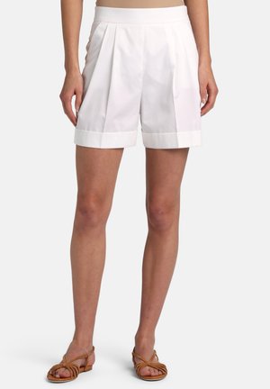 Frau trägt weiße, plissierte Shorts mit hohem Bund und umgeschlagenen Säumen sowie braune, flache, schmale Sandalen, steht vor einfarbigem Hintergrund.