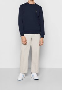 Námořnický svetr s žebrovanými manžetami a výstřihem, se malým zeleným logem. V kombinaci se světle béžovými kalhotami a bílými teniskami s modrými akcenty.