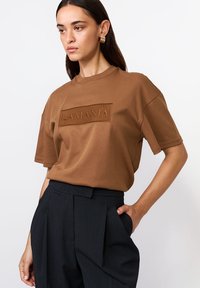 Kobieta w brązowej, oversize'owej koszulce z napisem "LAMANIA" oraz czarnych, wysokich, plisowanych spodniach, pozująca z jedną ręką w kieszeni.