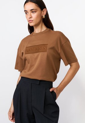 Kobieta w brązowej, oversize'owej koszulce z napisem "LAMANIA" oraz czarnych, wysokich, plisowanych spodniach, pozująca z jedną ręką w kieszeni.