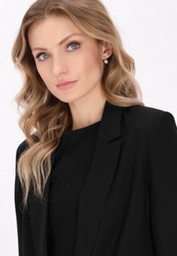 Černý blazer se špičatými klopy, nošený přes černý top; má vlnité světlé hnědé vlasy a perlovo náušnice. Jednoduché bílé pozadí.