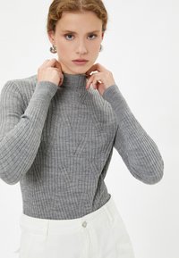 Maglione a coste grigio con colletto alto e motivi diagonali. Maniche lunghe con polsini aderenti. Abbinato a pantaloni bianchi a vita alta.