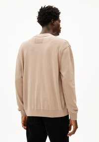 Beige gebreide trui met lange mouwen, een ronde hals en een klein merklabel op de achterkant. Gekozen in combinatie met zwarte corduroy broeken.