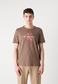 Pruun puuvillane T-särk, millel on roosa graafiline logo, mis ütleb "Calvin Klein Jeans" ja lühikesed varrukad, kantud beežide pükstega.