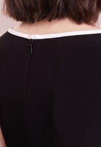 Svart klänning med vit halsringning, med dold dragkedja bak och en strukturerad tygkvalitet. Designen är stilren och figurnära.