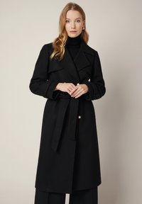 Czarny wełniany płaszcz trench z szerokimi klapami, paskiem w talii i złotymi guzikami. Posiada długie rękawy i lekko rozkloszowany dół.