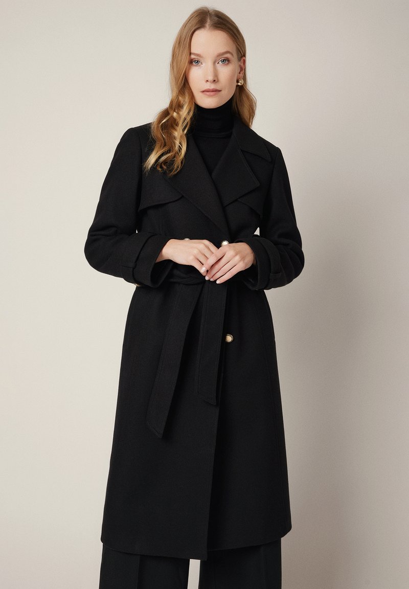 Czarny wełniany płaszcz trench z szerokimi klapami, paskiem w talii i złotymi guzikami. Posiada długie rękawy i lekko rozkloszowany dół.