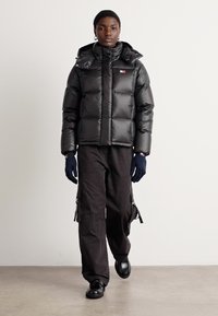 Juodas pūkuotas chalatas su tekstūruotu paviršiumi, aukštu apykakle ir užtrauktukais užsegamomis kišenėmis; derinamas su laisvomis juodomis kelnėmis, turinčiomis šoninius užtrauktukus, ir pirštinėmis.